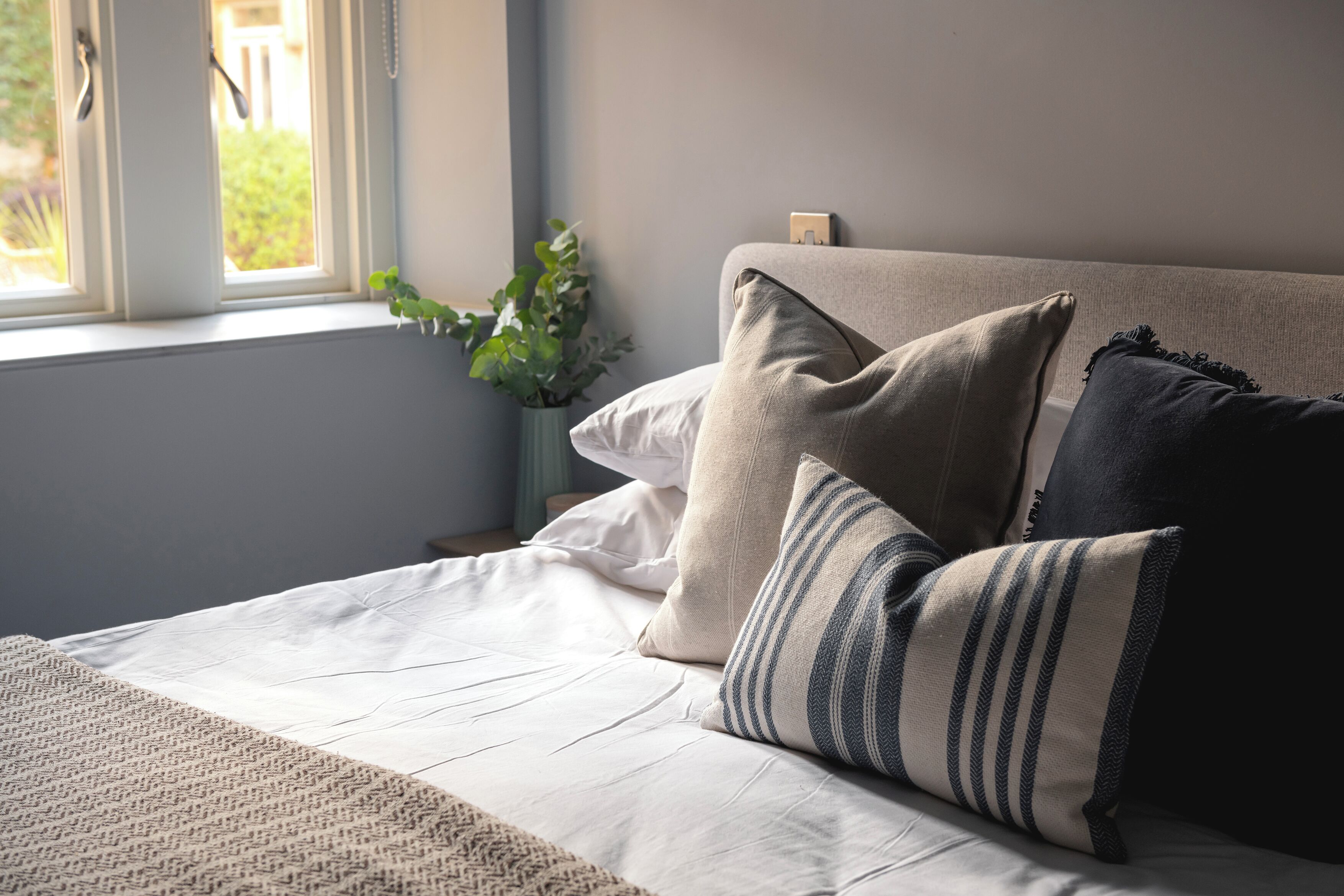 A neatly made bed with a beige and white duvet, a striped pillow, and a dark pillow. A small plant is visible in the corner.