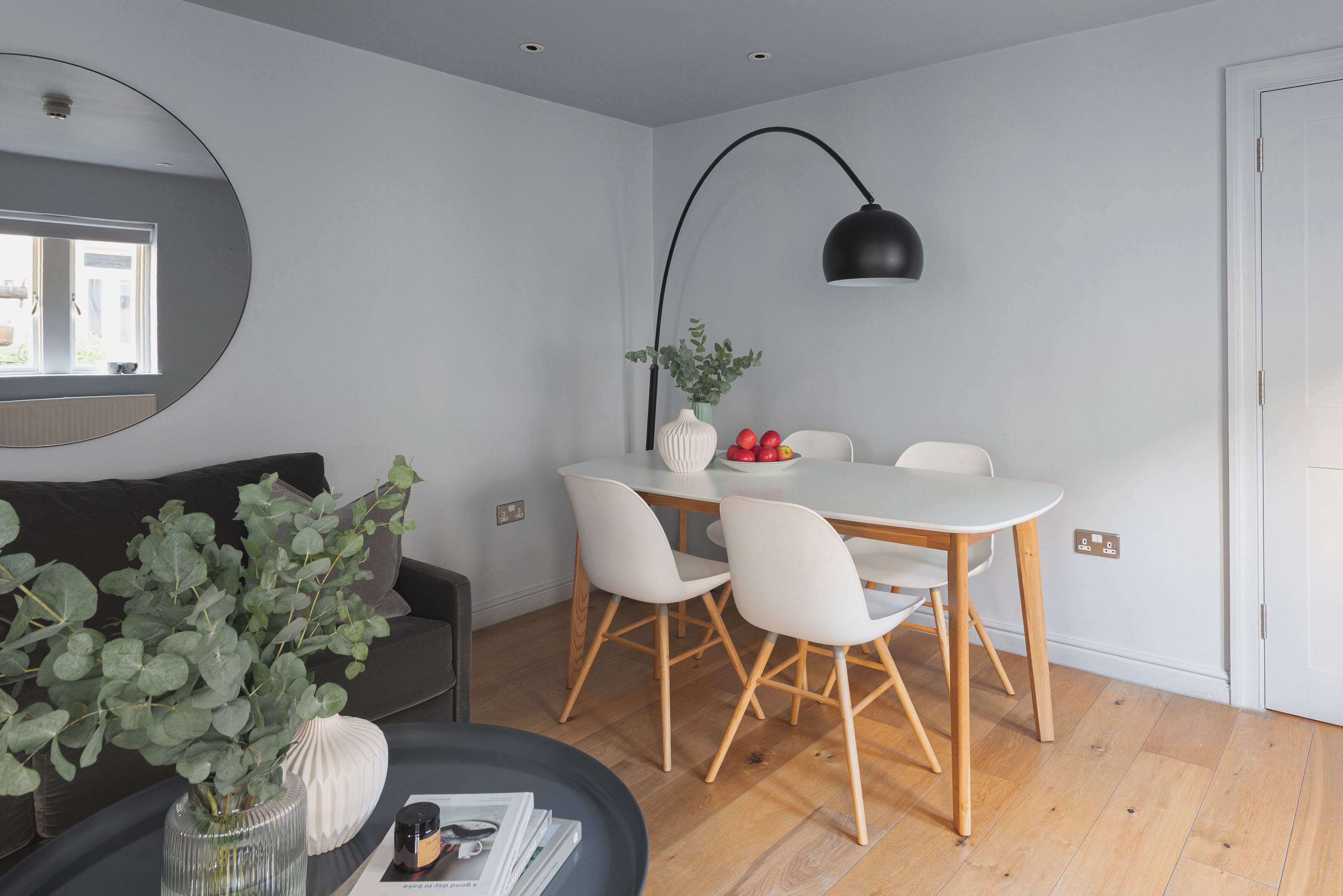 A modern living room with a round mirror, a dining table with chairs, a black floor lamp, and a potted plant.