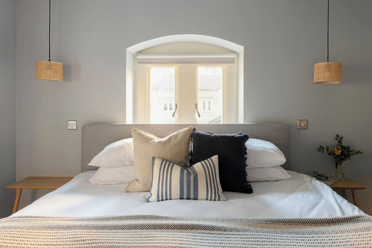 A neatly made bed with a patterned bedspread, a window with a view of buildings, and two hanging wicker lamps.