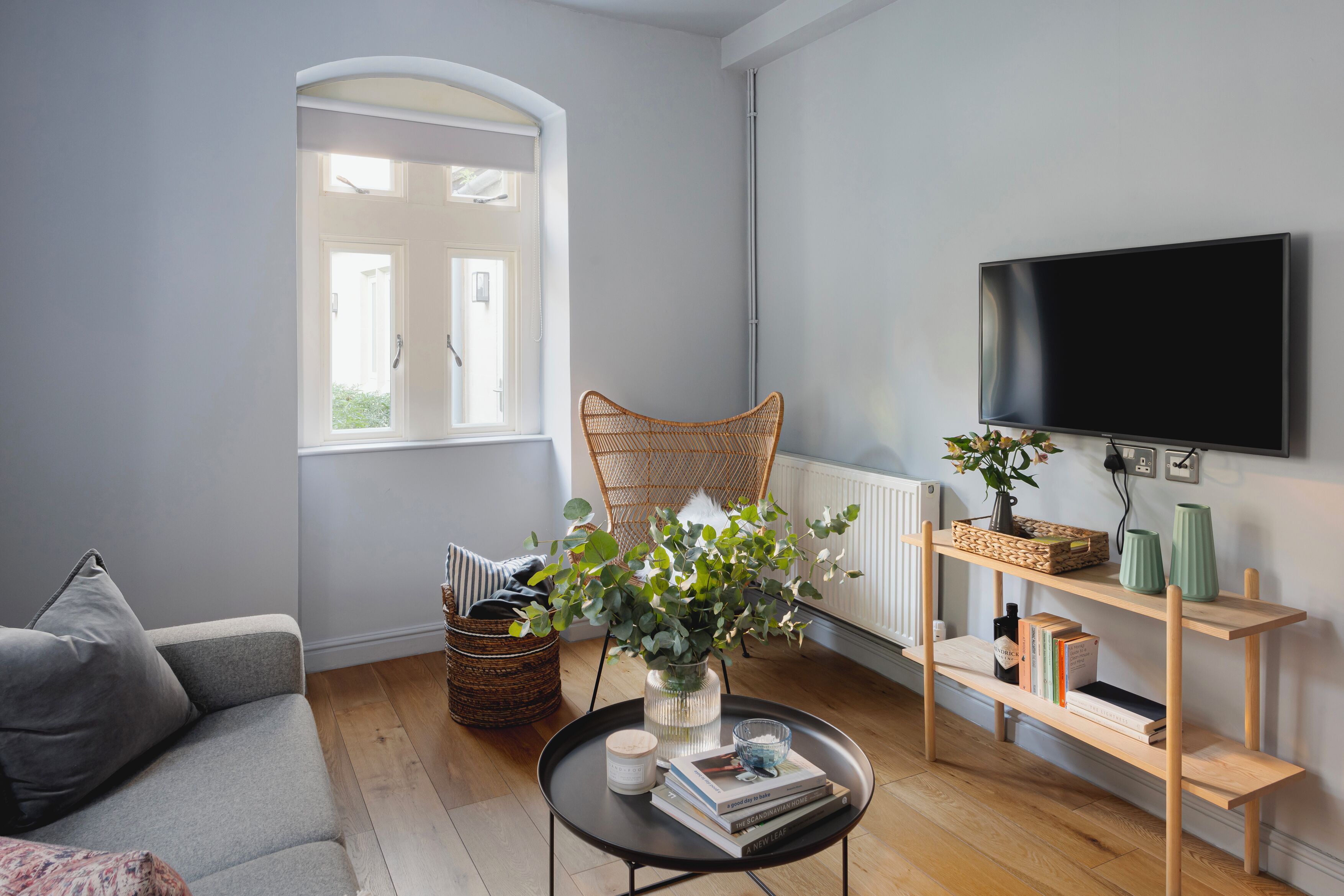 Un salon avec un canapé gris, une table basse ronde ornée de livres et d’un vase, un fauteuil en osier, une bibliothèque et un téléviseur fixé au mur.