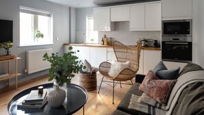 A modern kitchen with white cabinets, a dining area with a wicker chair, and a living room with a sofa and a coffee table. - St Catherine's Hospital - Bath (Bath)