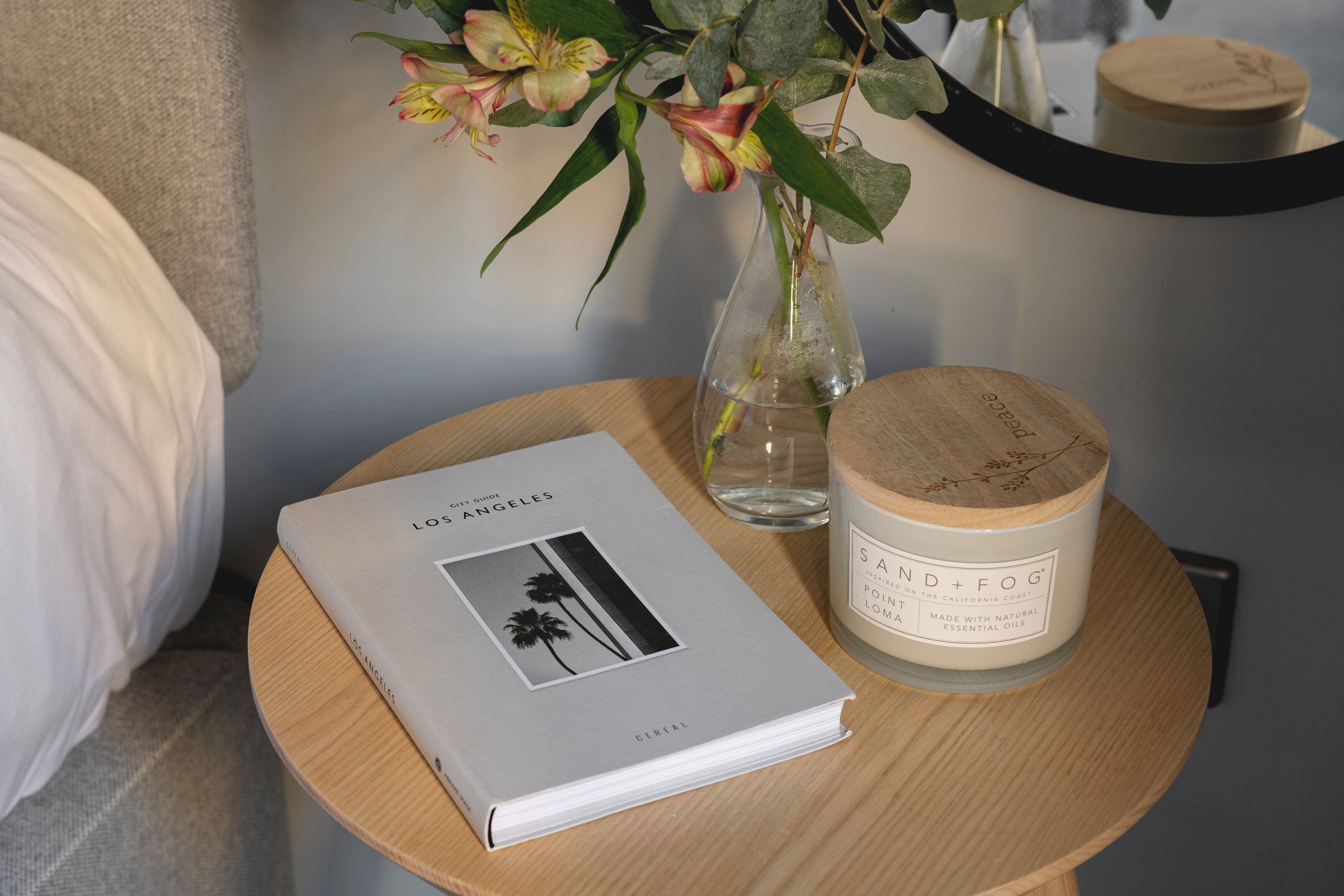 A book titled "Los Angeles" on a round wooden table, a candle with a wooden lid, and a vase with flowers.