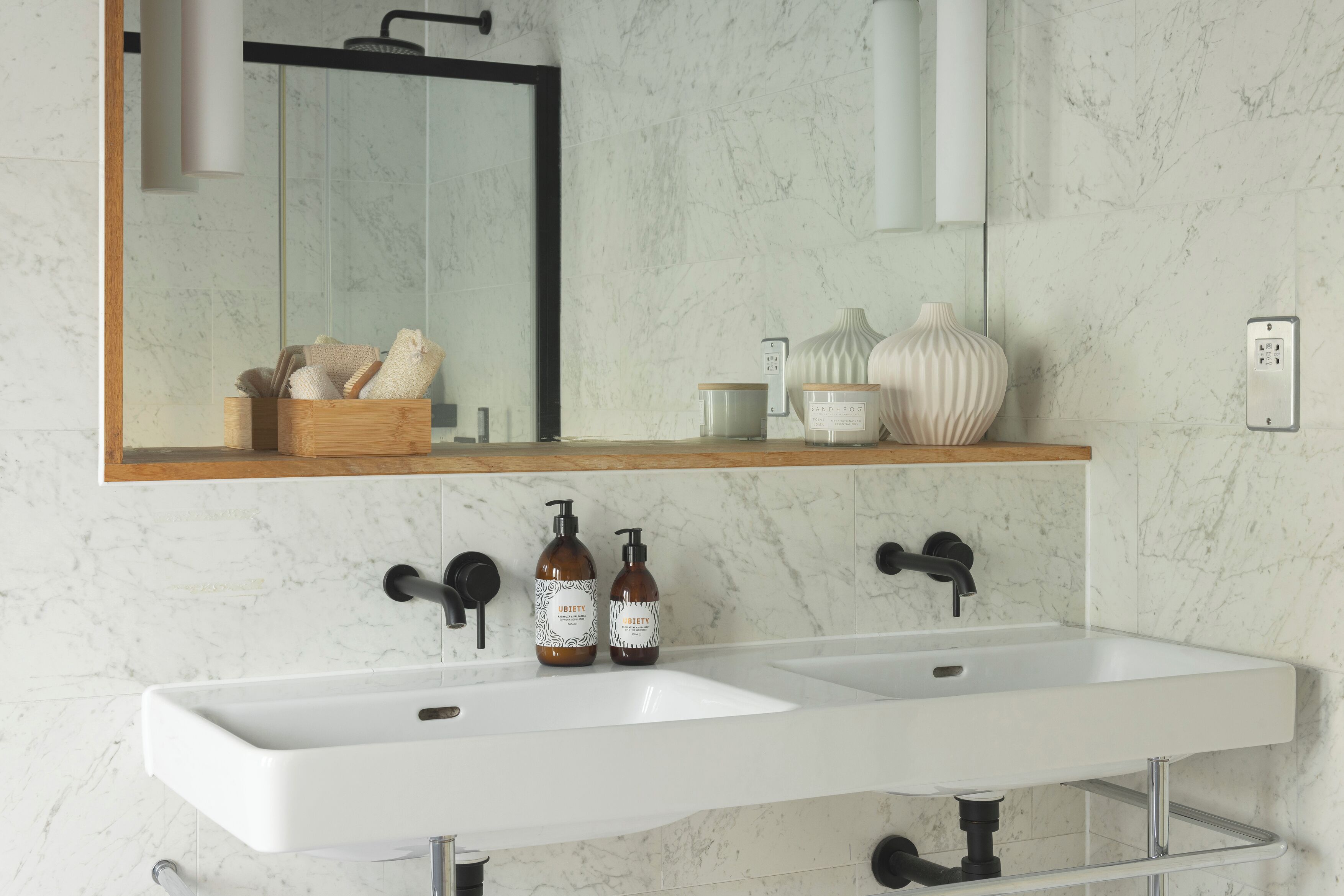 Un baño con un lavabo blanco, dos grifos negros y un espejo encima. Hay dos botellas de jabón en el lavabo, un jarrón pequeño y un caja de madera con toallas.