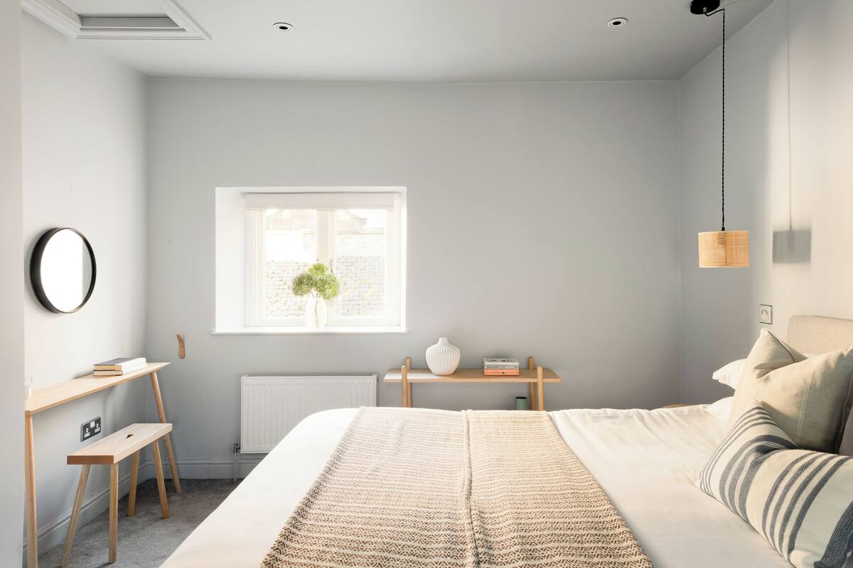 A bedroom with a bed, bedside table, desk, radiator, window, and hanging light fixture.