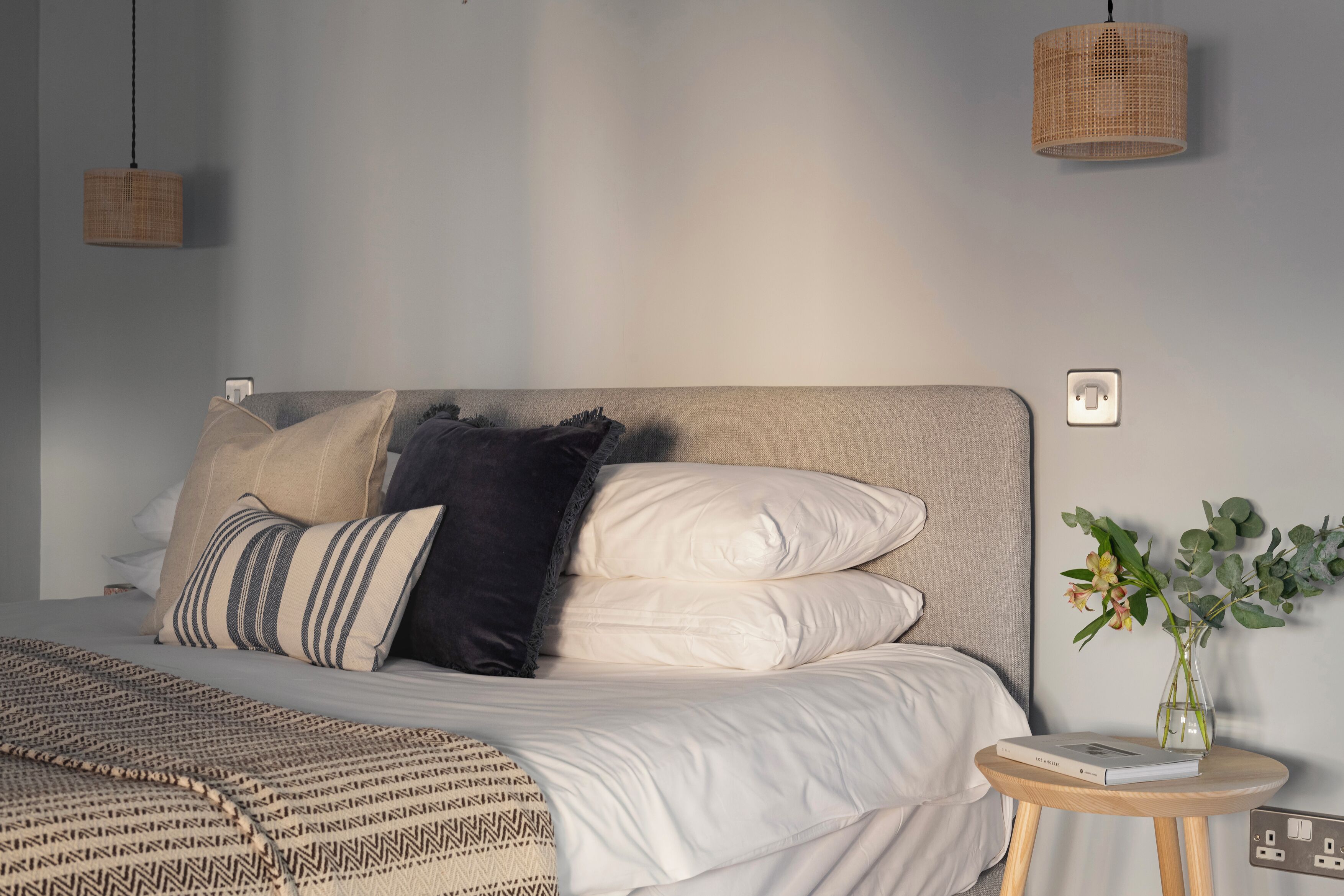 A neatly made bed with a patterned blanket, a bedside table with a book and a vase of flowers, and a hanging light fixture.
