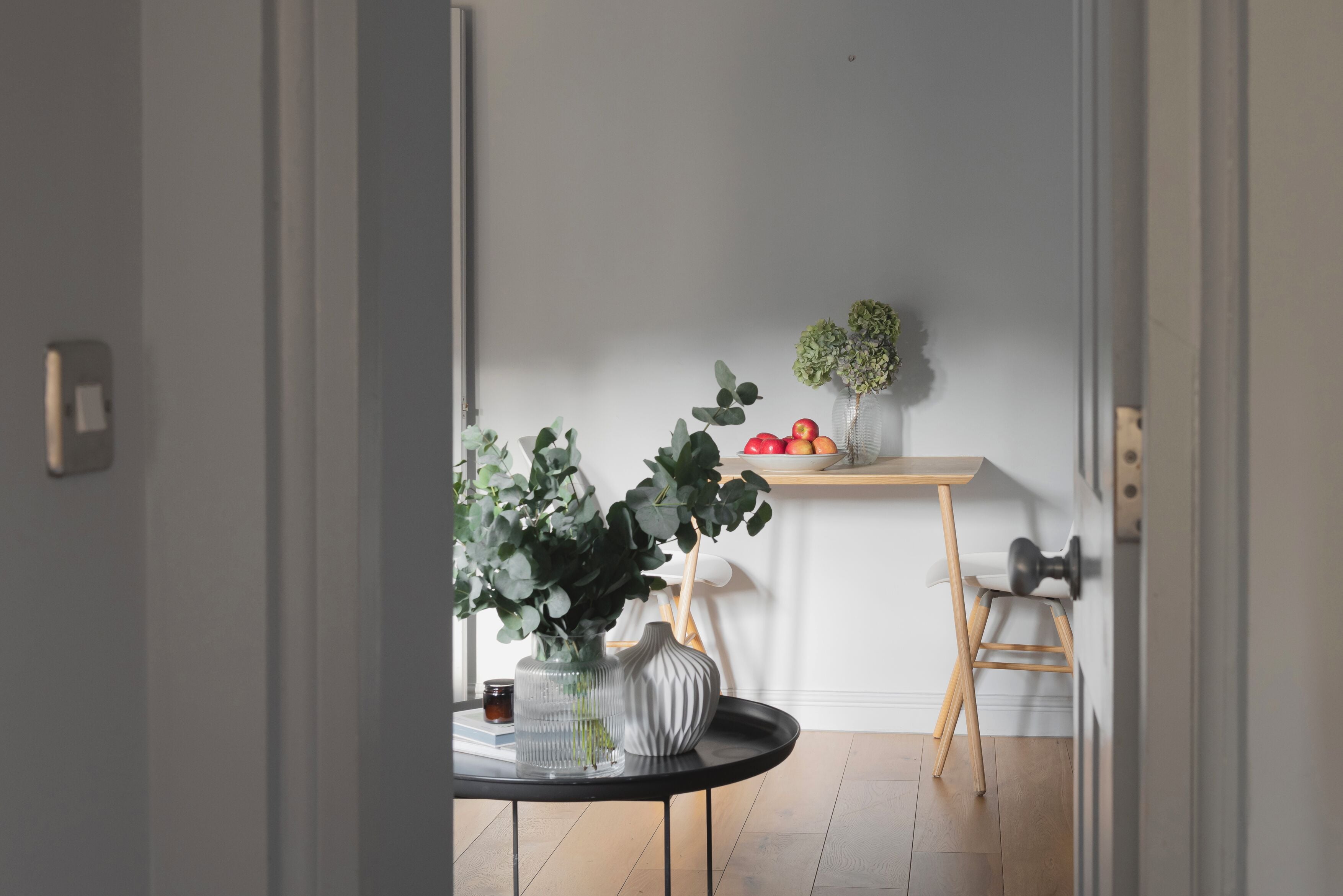A room with a round table, a small stool, a potted plant, and a shelf with apples.