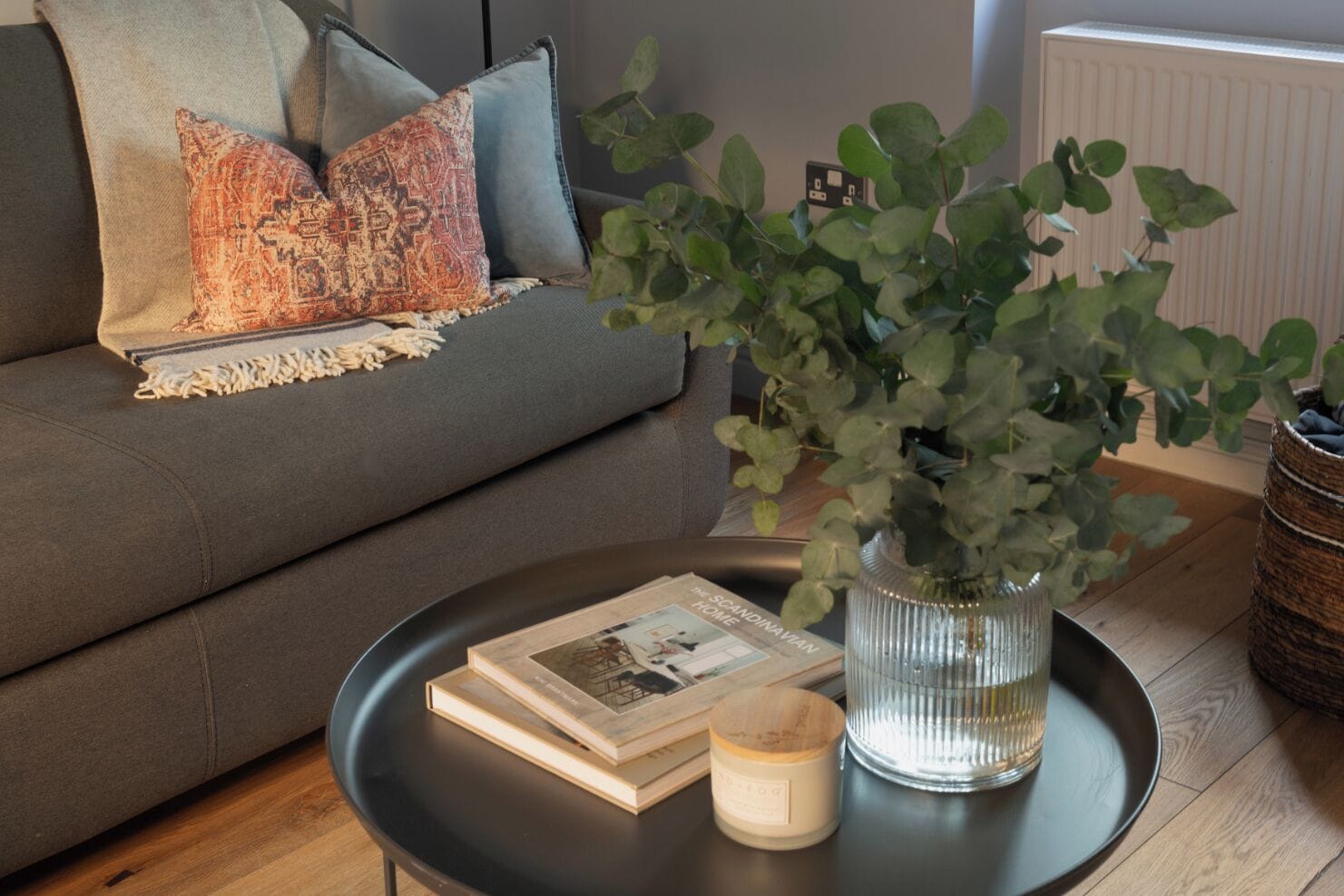 Ein Wohnzimmer mit grauem Sofa, einem schwarzen Couchtisch, einem Buch, einer Kerze und einer Pflanze in einer Vase.