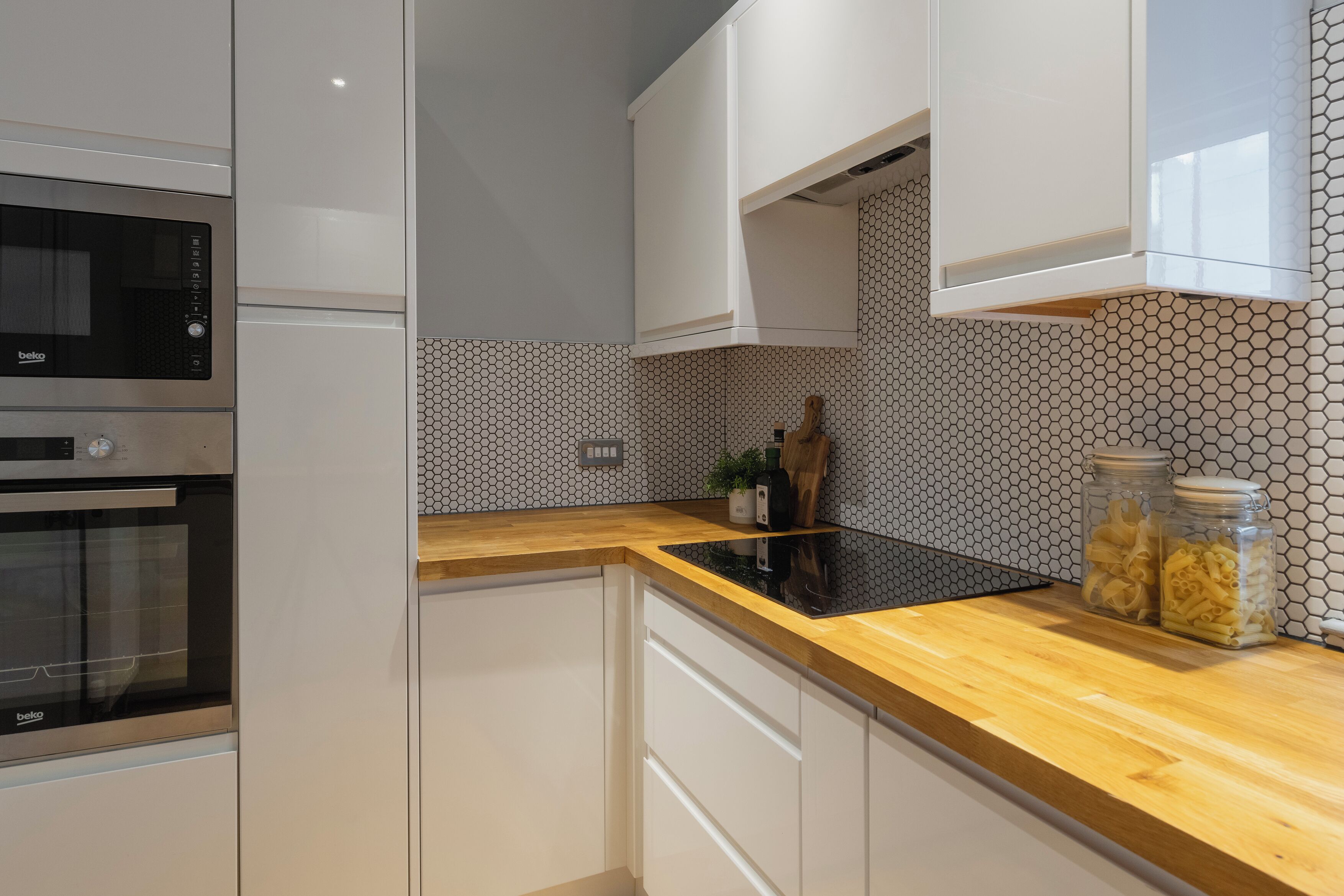 A kitchen with white cabinets, a wooden countertop, a black induction cooktop, and a microwave. There are two jars of pasta on the counter.