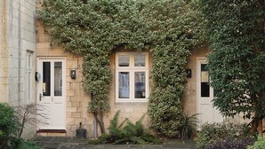 A stone building with a white door and windows, surrounded by lush greenery and a stone pathway. - St Catherine's Hospital - Bath (Bath)