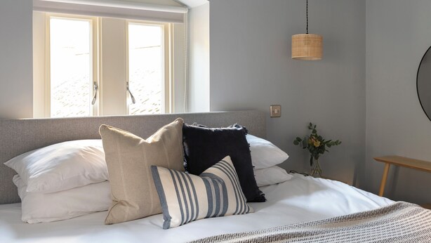 A neatly made bed with multiple pillows, a hanging light fixture, a window with a view, and a small table with a vase. - St Catherine's Hospital - Bath (Bath)