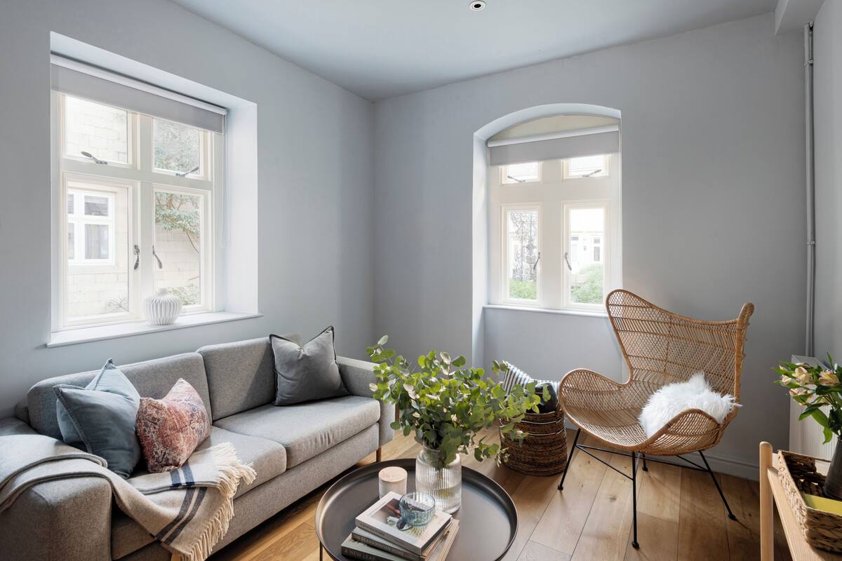 A living room with a sofa, armchair, and coffee table. There are windows with views of greenery and a brick wall.