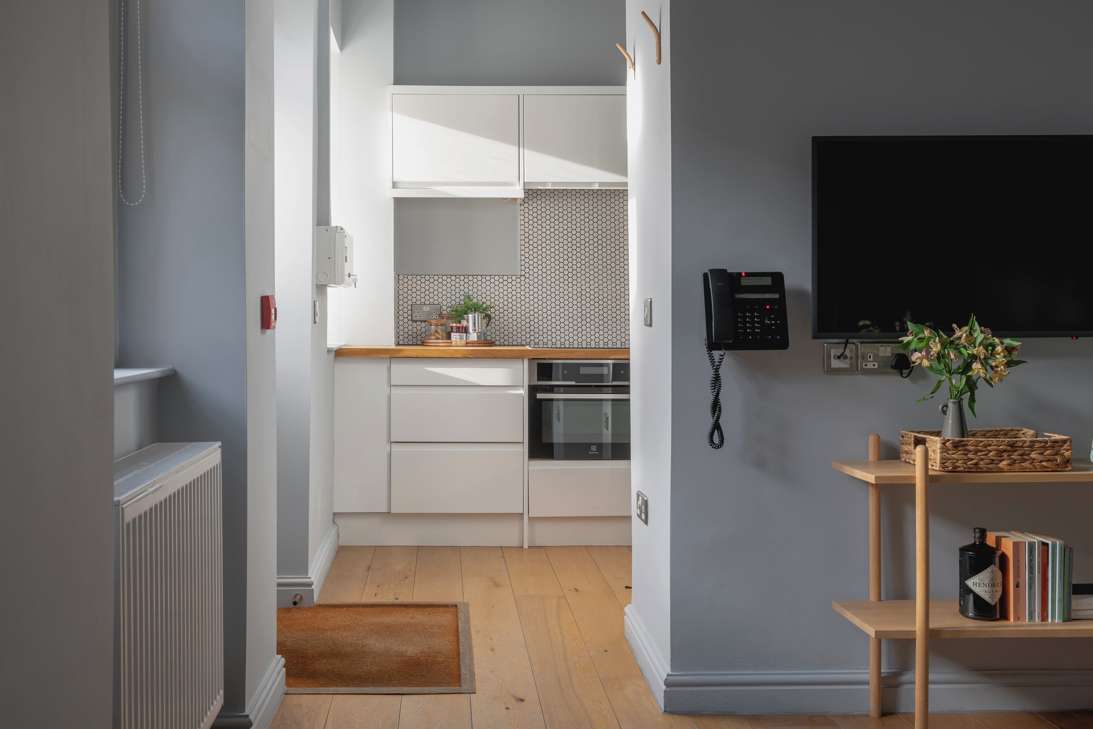 Una cocina moderna con armarios blancos, horno empotrado y microondas. Hay piso de madera, un tapete pequeño y una estantería con libros y un jarrón con flores.