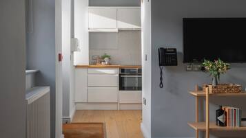 Una cocina moderna con armarios blancos, horno empotrado y microondas. Hay piso de madera, un tapete pequeño y una estantería con libros y un jarrón con flores.