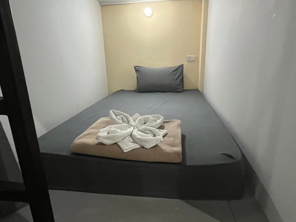 A small, single-bed room with a grey mattress, a beige pillow, and a towel arranged on the bed.