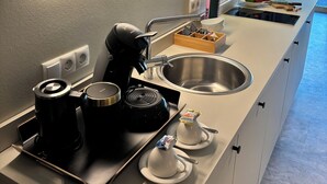 Cocina moderna equipada con cafetera, hervidor y microondas. Hay un lavamanos con pileta de acero inoxidable y una pequeña encimera con fruteros y una caja de pañuelos.