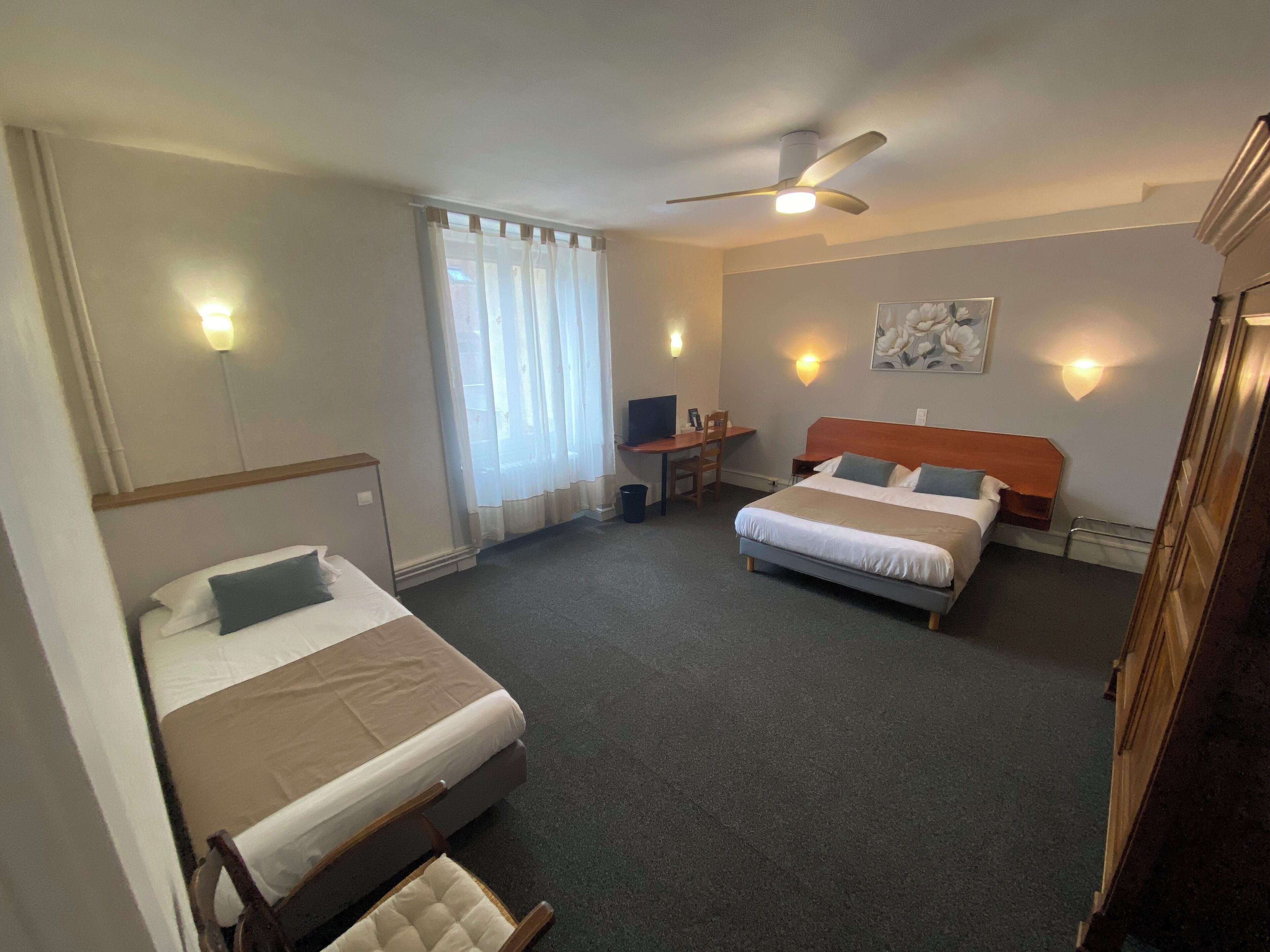 Une chambre d’hôtel avec deux lits, un bureau, une chaise, un ventilateur de plafond et une fenêtre avec des rideaux.