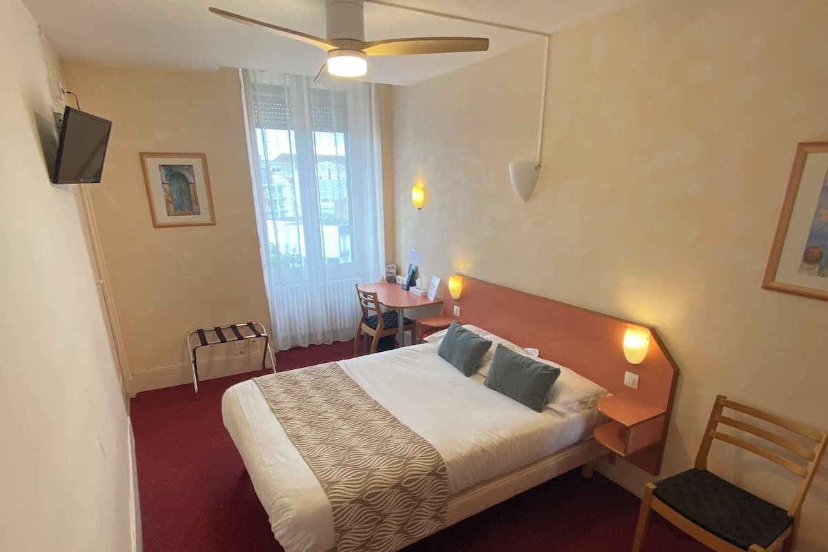 Une chambre d’hôtel comprenant un lit, une chaise, un bureau avec une chaise, un ventilateur de plafond et une fenêtre avec des rideaux.