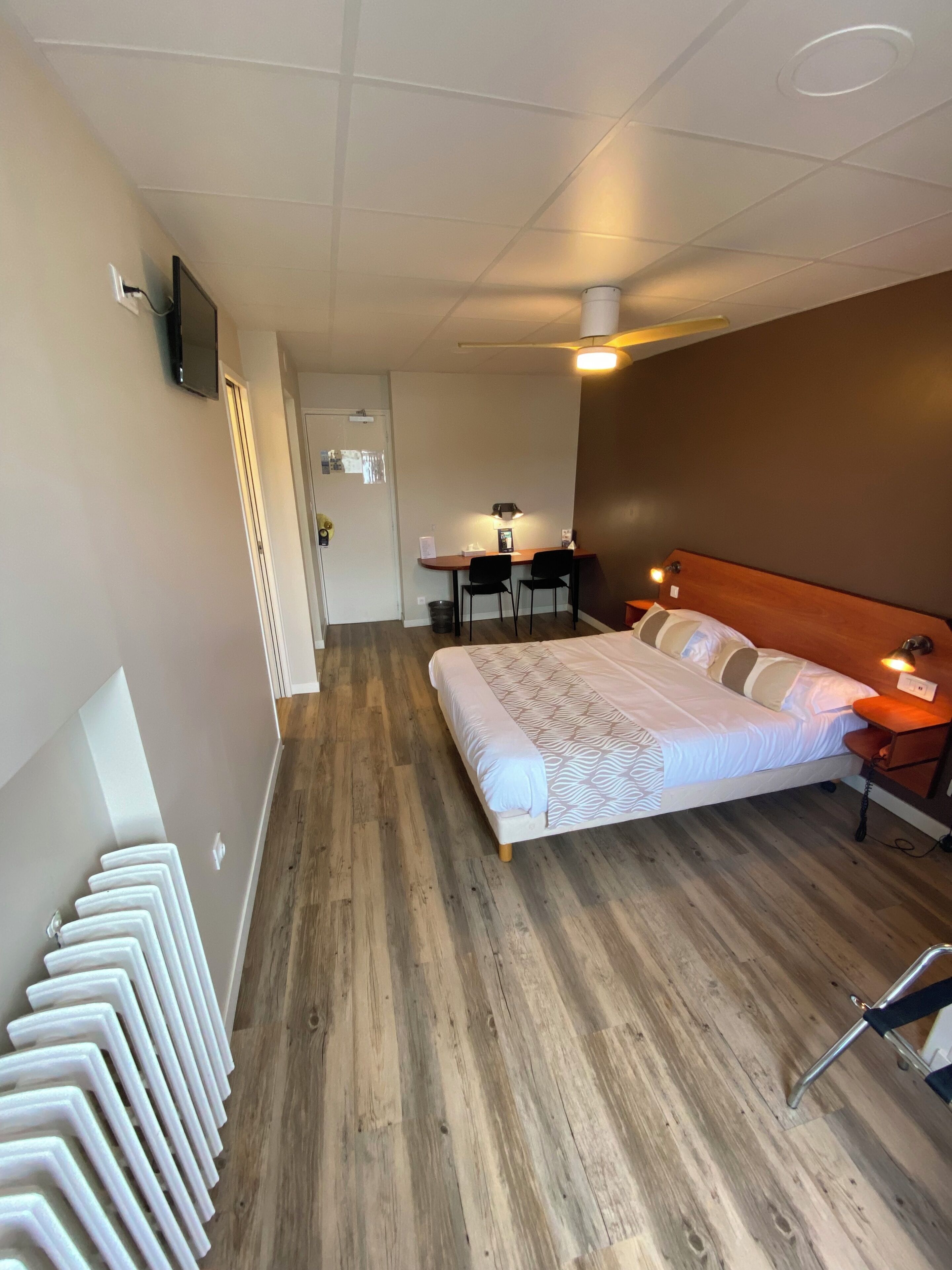 A hotel room with a bed, a desk with two chairs, a ceiling fan, and a TV mounted on the wall.