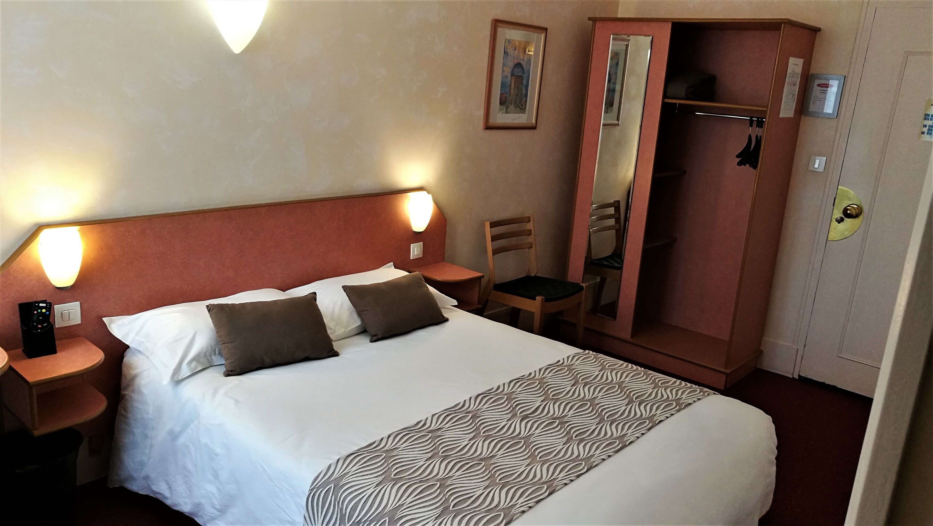 A hotel room with a bed, two bedside tables, a chair, a wardrobe, and a wall-mounted light fixture.