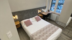 A modern bedroom with a bed, bedside tables, a desk, and a window with a view of parked cars. - NOUVEL HOTEL  (Lons-le-Saunier)