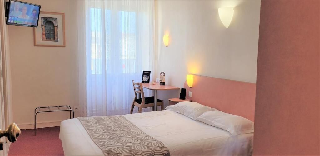 Une chambre d’hôtel avec un lit, une petite table et une chaise, un téléviseur fixé au mur et une fenêtre avec des rideaux légers.