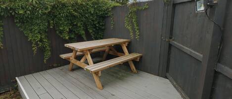 Une table de pique-nique en bois, avec des bancs, est posée sur une terrasse en bois, le tout étant entouré d’une clôture en bois sombre, ornée de plantes grimpantes.