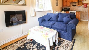 Una sala de estar con un sofá azul, una mesita con mantel floreado, una chimenea y una cocina al fondo.