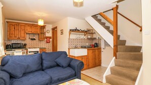Una sala de estar con un sofá azul, un mantel con estampado floral, y una cocina al fondo con armarios de madera y una escalera.