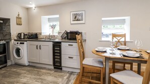 Cocina moderna con lavadora integrada, microondas y un lavamanos. Hay una zona de comedor con una mesa preparada para dos personas y un ventanal que proporciona luz natural.