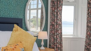 A hotel room with a bed, a nightstand, a lamp, a window with curtains, and a view of a landscape. - Llys Owen (Criccieth)