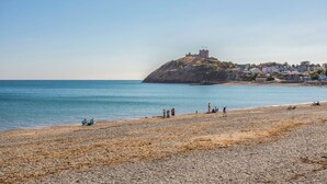 A beach with pebbles and a castle on a hill in the background. - Llys Owen (Criccieth)