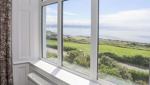 A room with large windows offering a view of a coastal landscape, including a road and greenery. - Llys Owen (Criccieth)