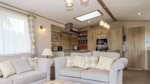 A modern living room with a sofa set, ottoman, and a kitchen area with wooden cabinets and appliances. - Ambleside (Cullompton)
