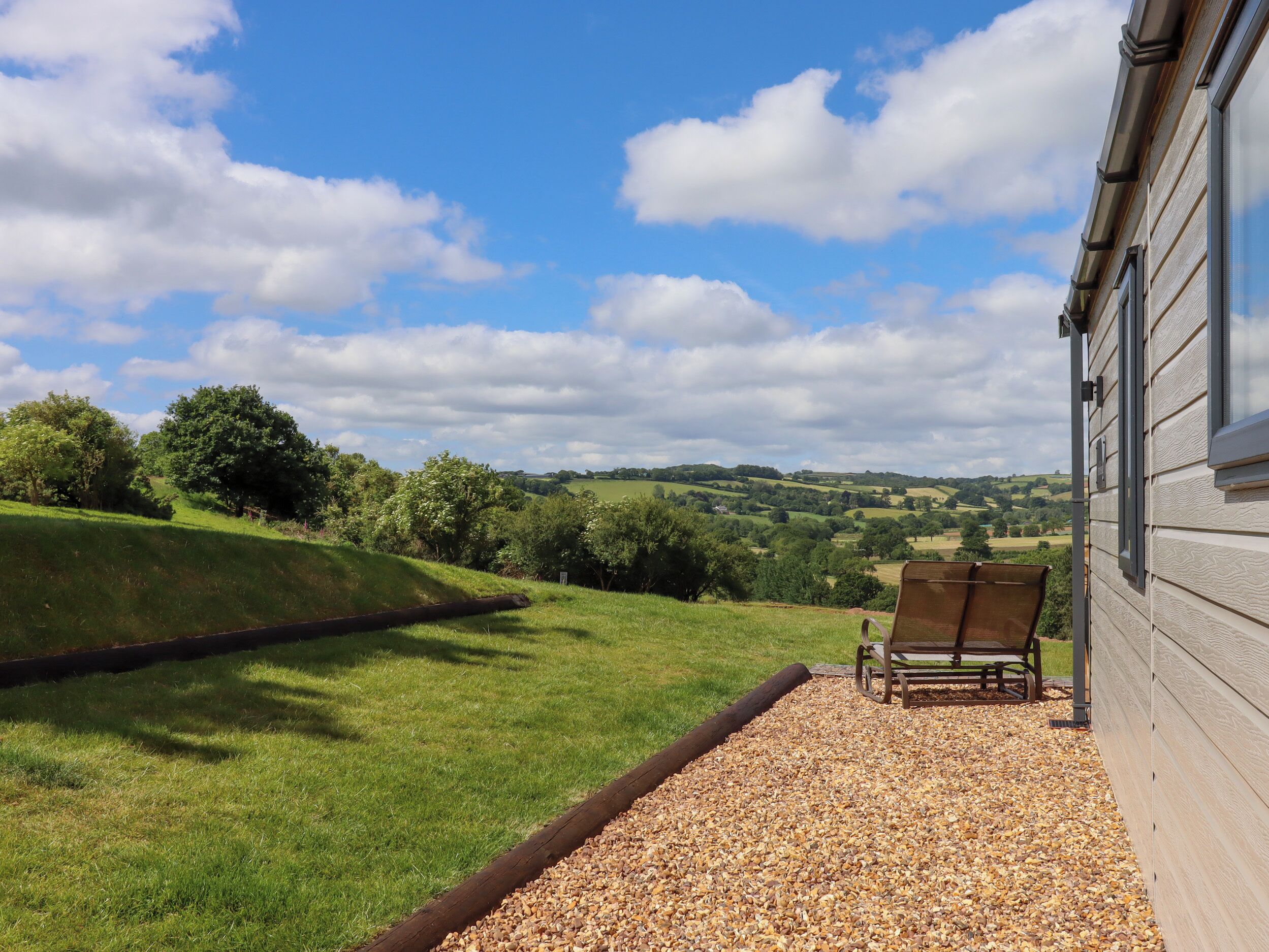 A patio with a gravel path, a wooden bench, and a view of rolling hills and trees under a partly cloudy sky.