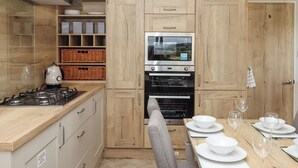 A modern kitchen with wooden cabinetry, a built-in oven, and a dining area set for four. - Ambleside (Cullompton)