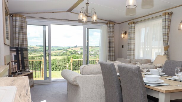 A modern living room with a balcony, a dining area with a table set for four, and a television in the corner. - Ambleside (Cullompton)