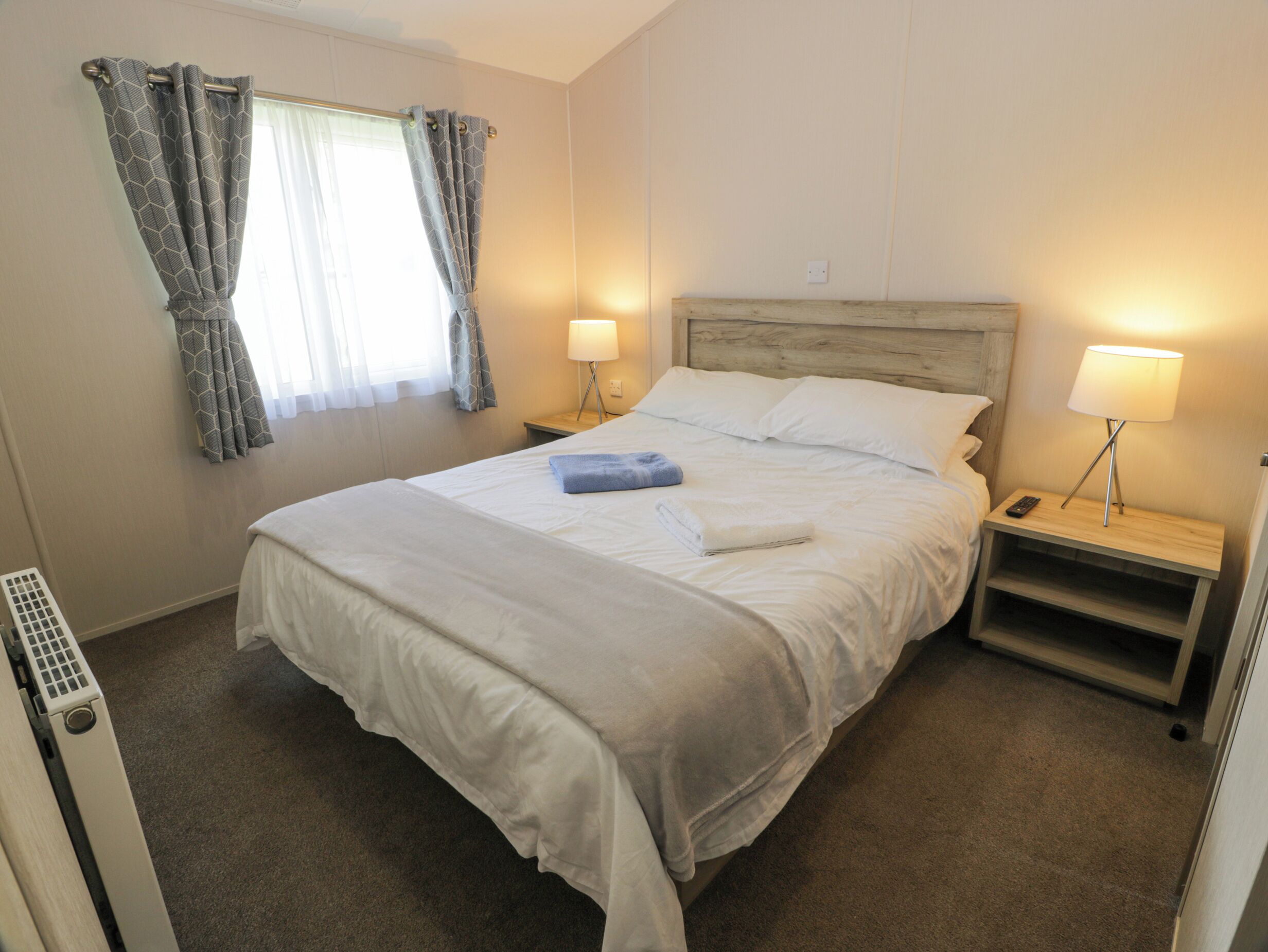 A neatly made bed with white linens, two bedside lamps, a nightstand, and a window with curtains.