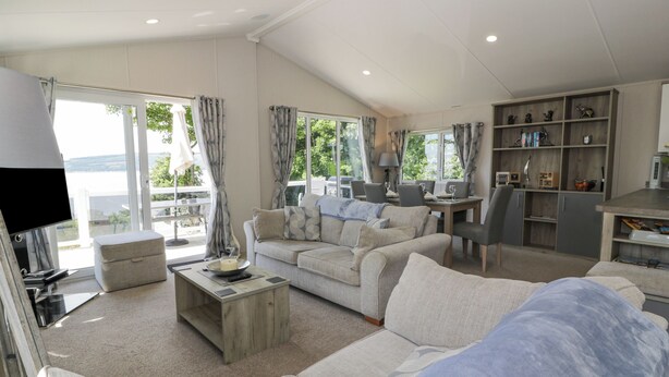 Un salon spacieux avec un canapé, une table basse et un téléviseur. Il y a également un coin repas avec une table et des chaises. La pièce est dotée de grandes fenêtres avec des rideaux et offre une vue sur l’extérieur.