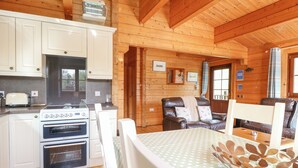 A wooden cabin interior with a kitchen area featuring white cabinets, a stove, and a toaster. There is a dining table with a polka dot tablecloth and a decorative centerpiece. - Heron Lodge (Bury St Edmunds)