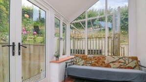 A conservatory with a bench, a small table, and a view of a garden. - Rose Cottage (Okehampton)
