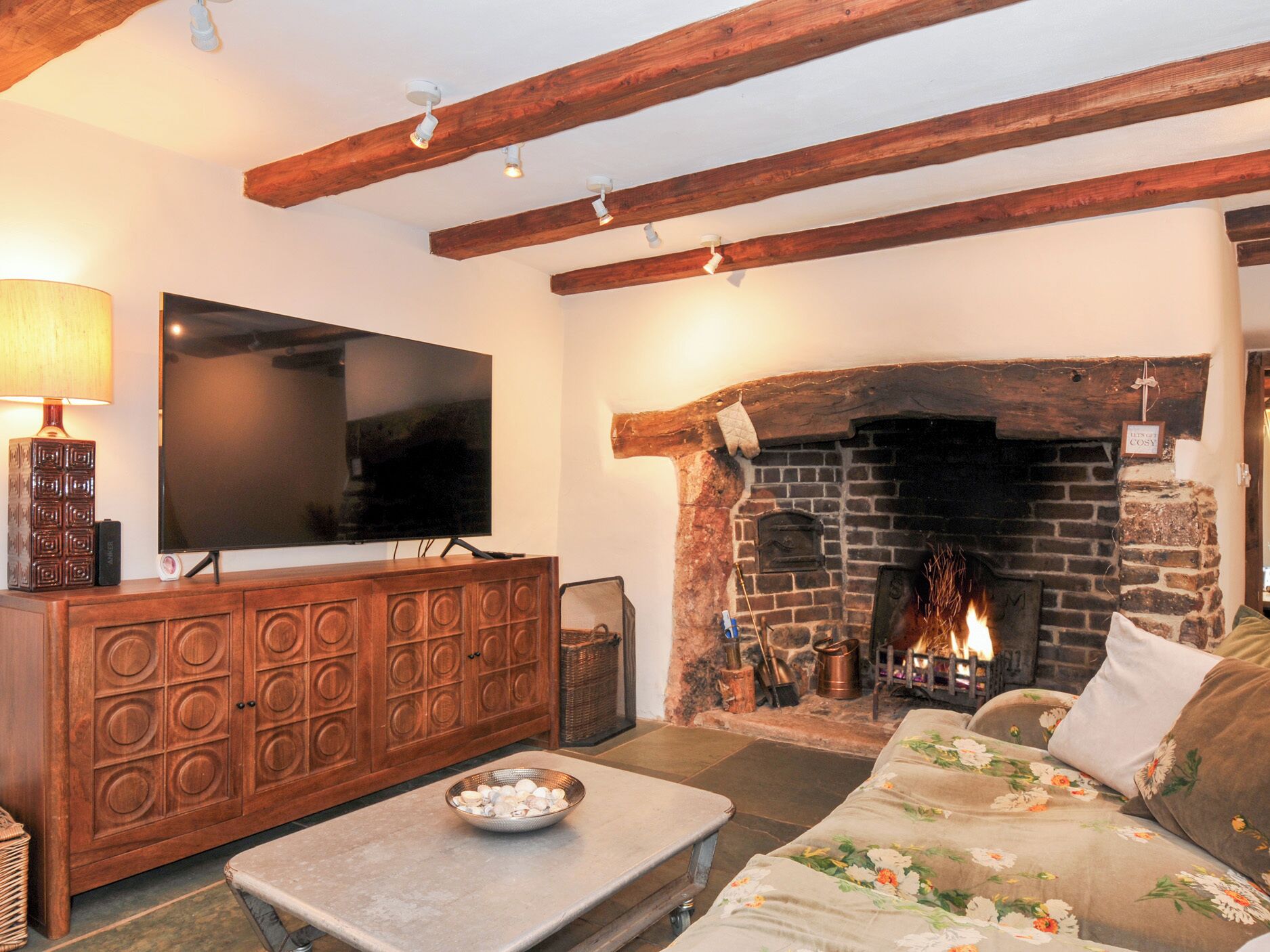 A cozy living room with exposed wooden beams, a flat-screen TV, a fireplace, and a floral sofa.