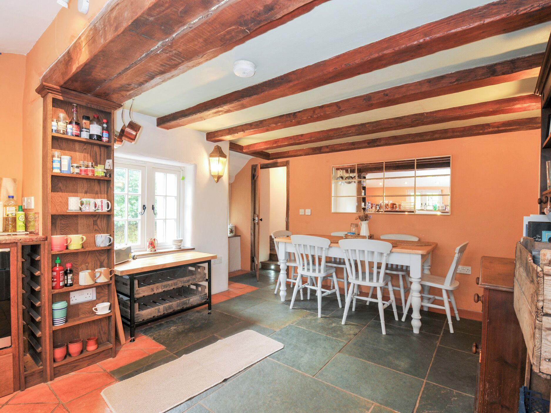 A rustic dining area with a wooden table and chairs, a built-in shelf with kitchen items, and a stone-tiled floor.