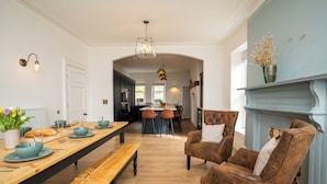 A dining area with a wooden table, benches, and chairs, a kitchen in the background, and a fireplace with a vase of flowers. - Haulfryn (Barmouth)