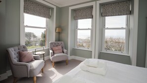 A bedroom with a large bed, two armchairs, a small table, and three windows with patterned valances. - Haulfryn (Barmouth)