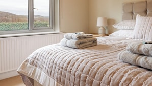 A bedroom with a bed, a window with a view, a bedside table with a lamp, and a stack of towels. - The Crib (Llandudno)
