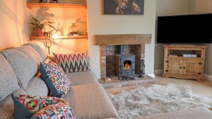 A cozy living room with a fireplace, a sofa with colorful cushions, a TV on a wooden stand, and a bookshelf with books and decorative items. - The Crib (Llandudno)
