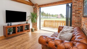 A living room with a leather couch, a TV on the wall, a wooden console table, and a large window with a view of a deck and trees. - Molly's Cottage (Malton)