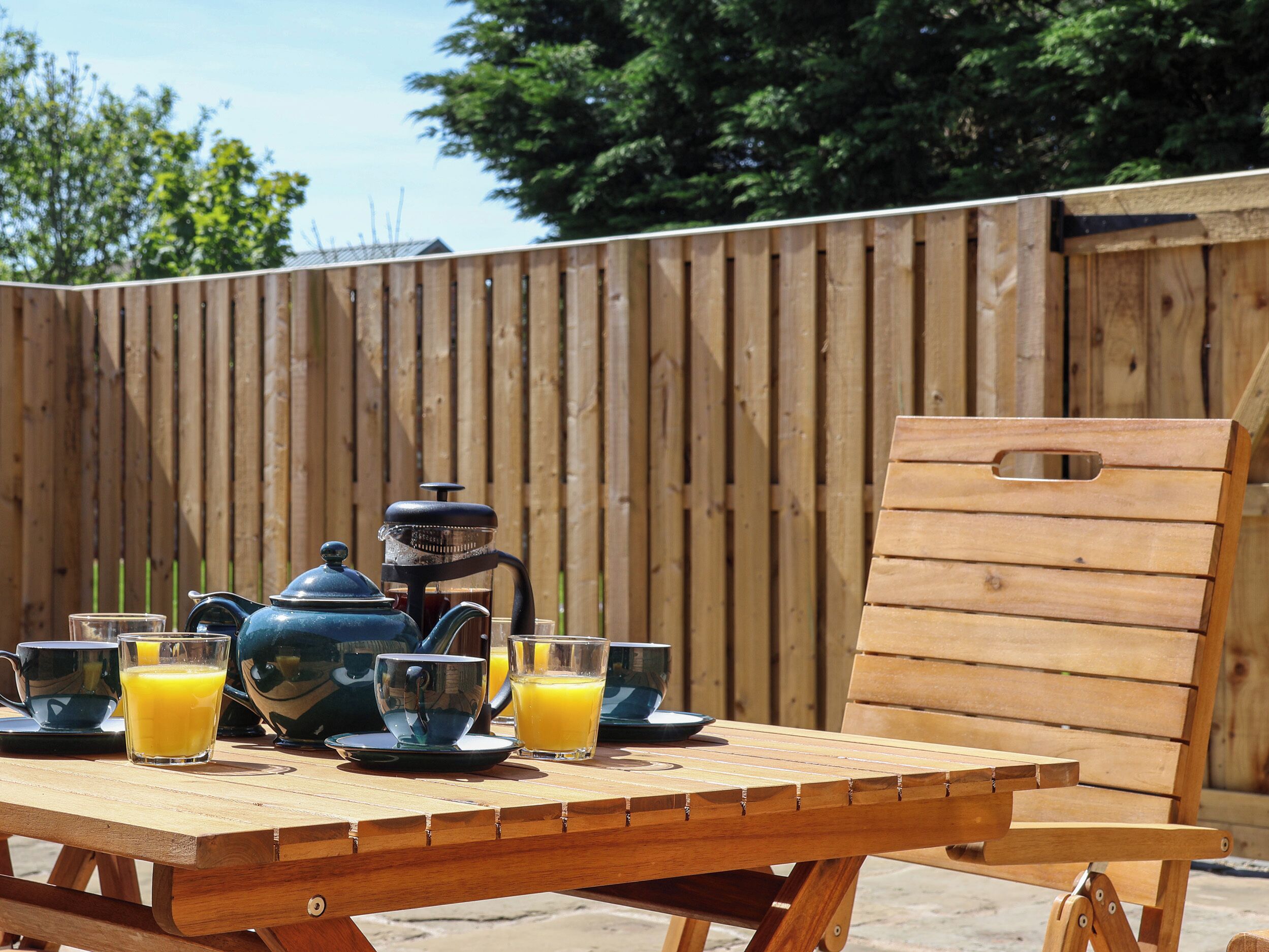 A wooden table with a French press, two glasses of orange juice, and a cup on a patio with a wooden fence in the background.
