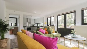 A modern living room with a mustard sofa, a green armchair, and a coffee table. There is a kitchen in the background with white cabinets and a window. - Cherry Orchard Farm (Maidstone)