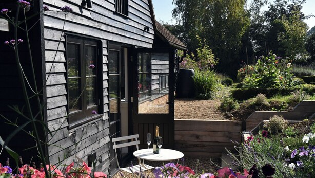 A wooden cabin with a small patio area, a white table with a bottle and glass, and a variety of colorful flowers in the foreground. - Cherry Orchard Farm (Maidstone)